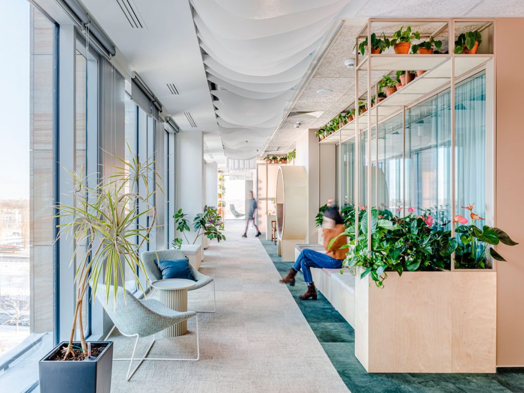 Jasna, szeroka aleja biurowa biegnąca wzdłuż szklanej ściany wieżowca, wypełniona naturalnym światłem. Po lewej stronie znajdują się minimalistyczne fotele wypoczynkowe, a po prawej wysokie, drewniane donice z gęstą roślinnością, które tworzą naturalną barierę dla strefy pracy. Pod sufitem ciągną się białe, faliste panele akustyczne, które nadają wnętrzu lekkości i spokoju. W tle widoczna jest postać w ruchu oraz charakterystyczny, okrągły mebel akustyczny.