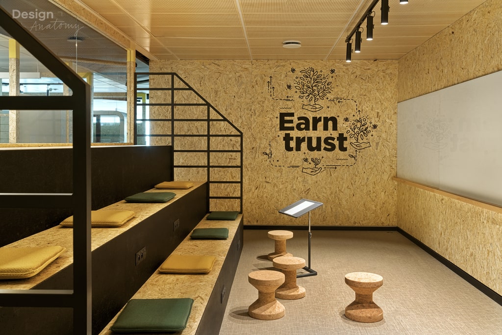 Wnętrze kreatywnej sali spotkań wyłożone płytami OSB na ścianach, z dużym napisem „Earn trust” i grafikami przedstawiającymi rośliny wyrastające z dłoni. W pomieszczeniu znajduje się czarna, schodkowa konstrukcja audytoryjna z żółtymi i zielonymi poduszkami do siedzenia oraz trzy korkowe taborety w kształcie klepsydry. Na ścianie po prawej stronie zamontowano dużą tablicę suchościeralną, a sufit kasetonowy z jasnego drewna posiada punktowe, czarne oświetlenie szynowe.