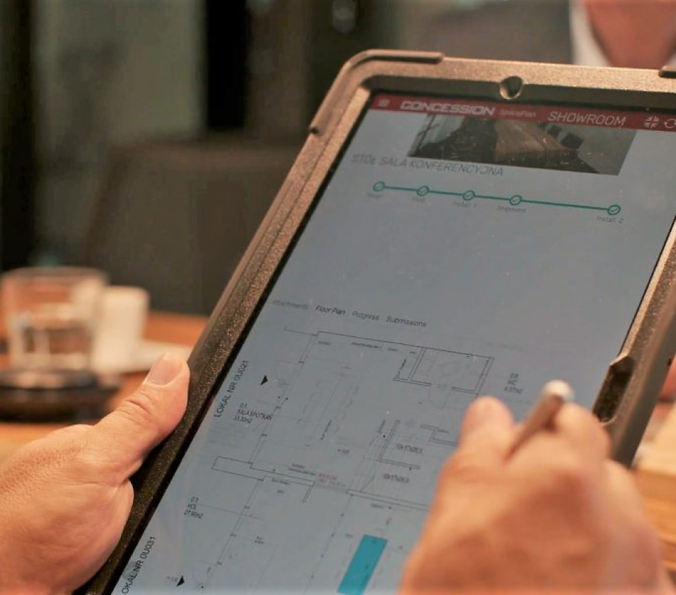 Zbliżenie na ręce trzymające tablet w pancernej obudowie, na którym wyświetlona jest aplikacja do planowania przestrzeni "CONCESSION SpacePlan". Na ekranie widoczny jest rzut techniczny pomieszczenia z zaznaczonymi meblami oraz pasek postępu realizacji projektu z etapami takimi jak: Design, Prod, Install 1, Shipment, Install 2. Osoba na zdjęciu posługuje się rysikiem, co sugeruje precyzyjne nanoszenie poprawek na planie.
